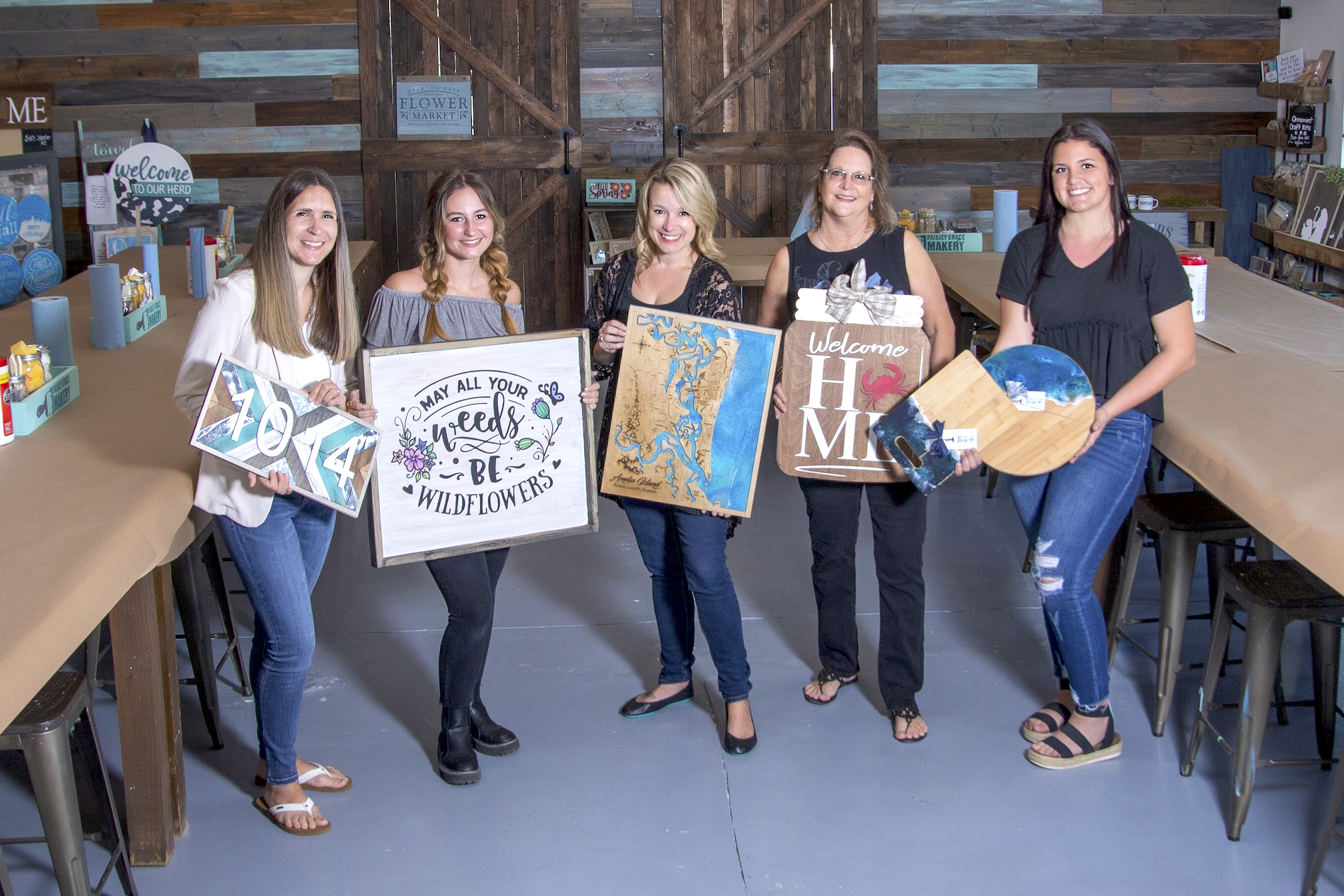 Five women stand in a craft studio, smiling and holding their finished art projects, including signs, paintings, and wood crafts. The background features wooden doors and tables with art supplies.