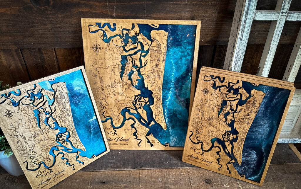 Three wooden maps featuring Amelia Island, Florida, are displayed against a rustic background. The maps show detailed engraved outlines and blue water accents, with each map varying in size and artistic shading.