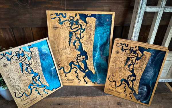 Three wooden maps featuring Amelia Island, Florida, are displayed against a rustic background. The maps show detailed engraved outlines and blue water accents, with each map varying in size and artistic shading.