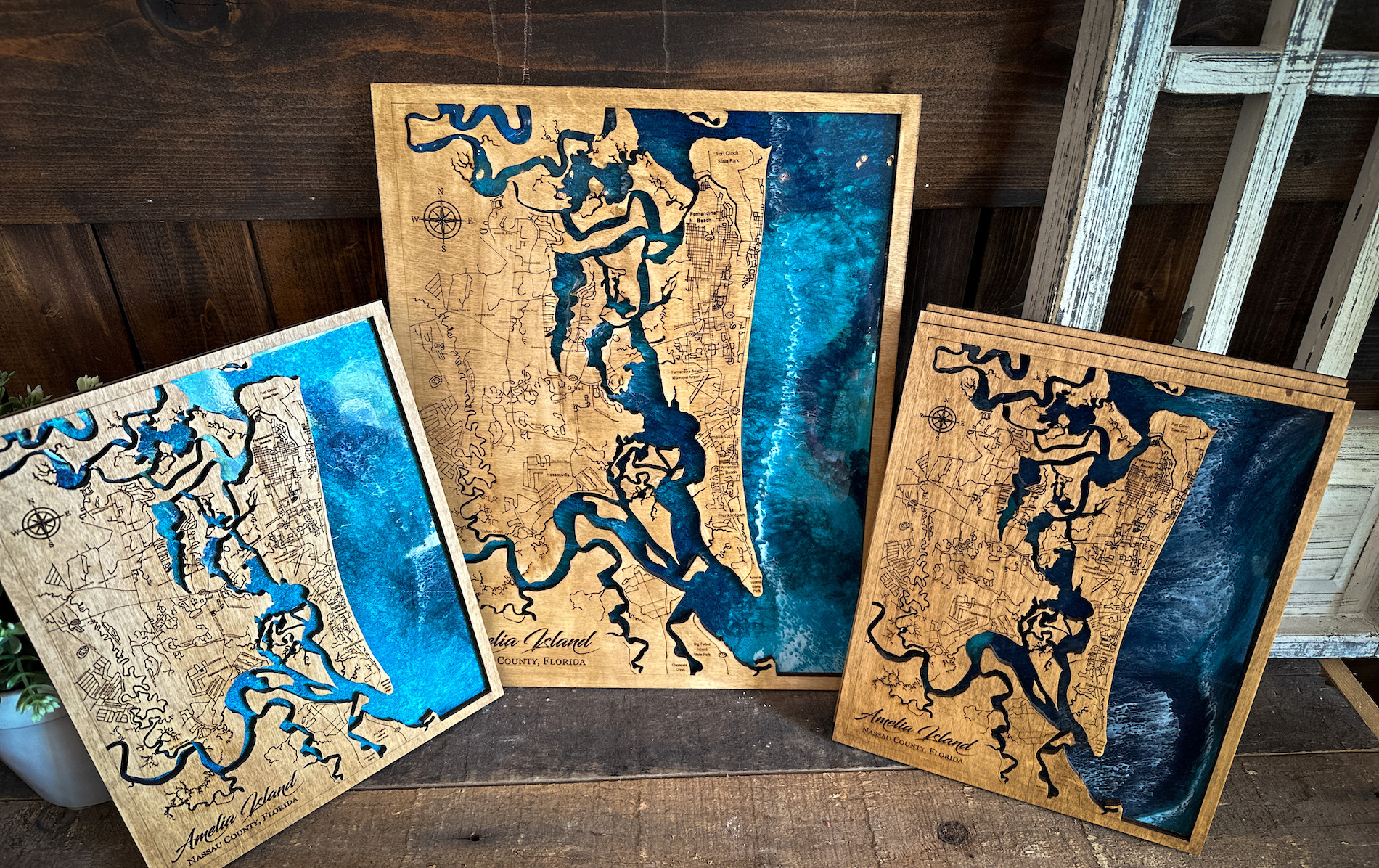 Three wooden maps featuring Amelia Island, Florida, are displayed against a rustic background. The maps show detailed engraved outlines and blue water accents, with each map varying in size and artistic shading.