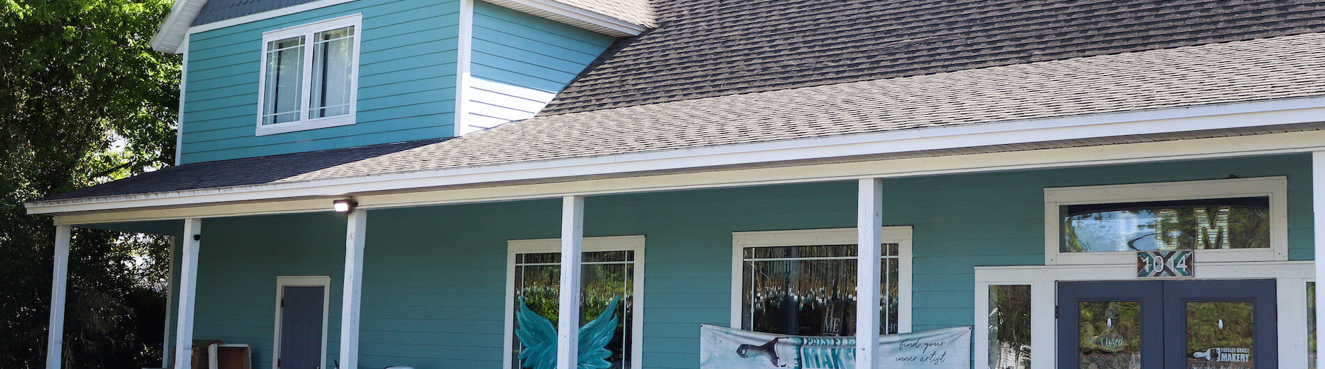 A turquoise, two-story building with a covered porch, white trim, and chairs outside. A painted mural of angel wings is visible between two large windows, and a palm tree stands behind the building.