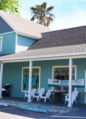 A turquoise, two-story building with a covered porch, white trim, and chairs outside. A painted mural of angel wings is visible between two large windows, and a palm tree stands behind the building.