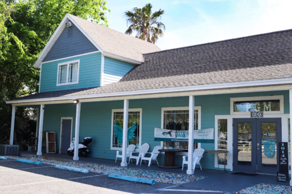 A turquoise, two-story building with a covered porch, white trim, and chairs outside. A painted mural of angel wings is visible between two large windows, and a palm tree stands behind the building.