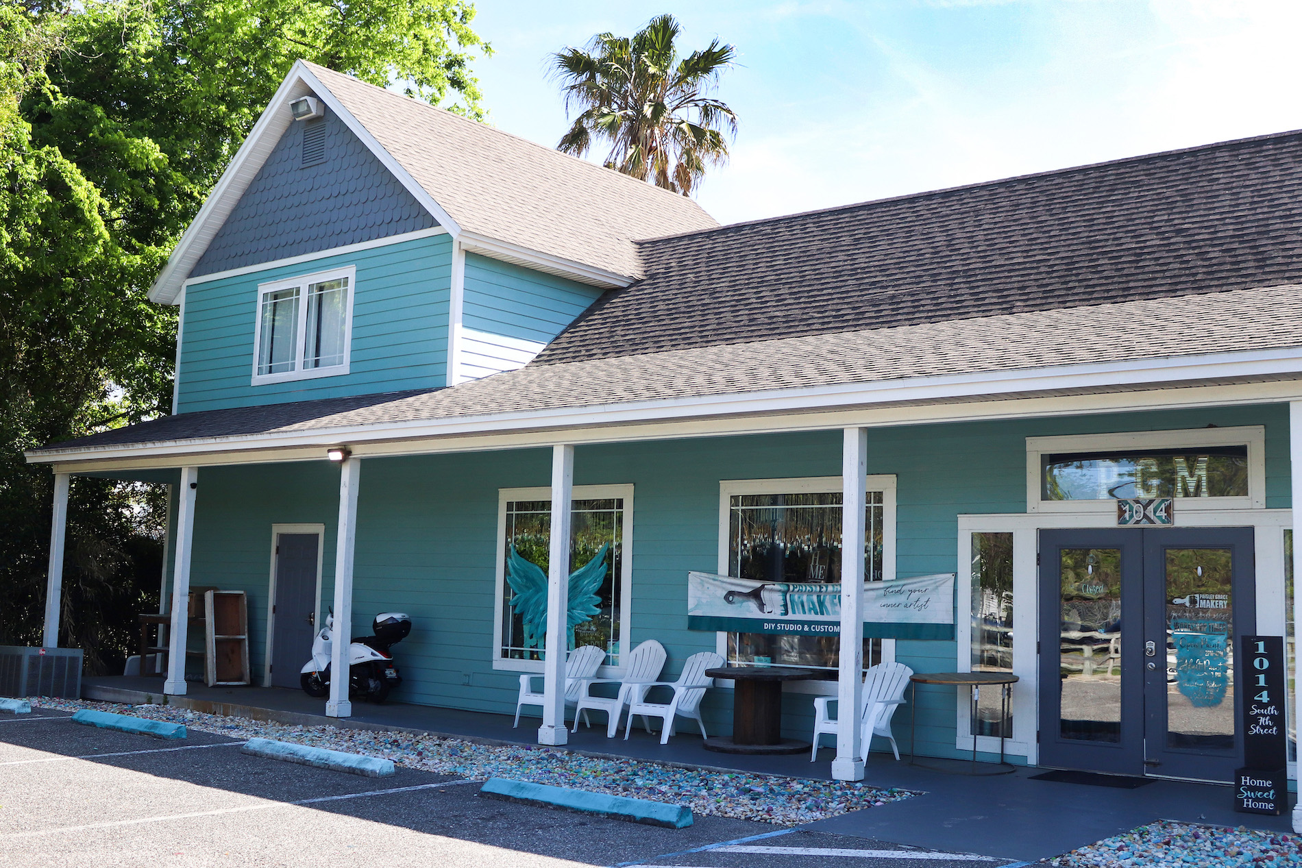 A turquoise, two-story building with a covered porch, white trim, and chairs outside. A painted mural of angel wings is visible between two large windows, and a palm tree stands behind the building.