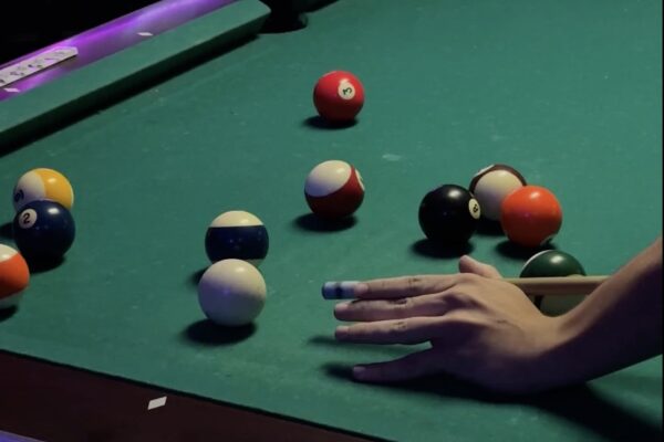 A hand poised with a cue stick prepares to take a shot on a green pool table, with several colorful billiard balls scattered across the surface.