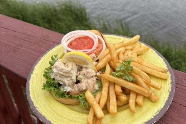 A plate with French fries and an open-faced seafood salad sandwich topped with lettuce, tomato, onion slices, and a lemon wedge at Palms Fish Camp, set on a wooden railing overlooking the water and marsh grass.