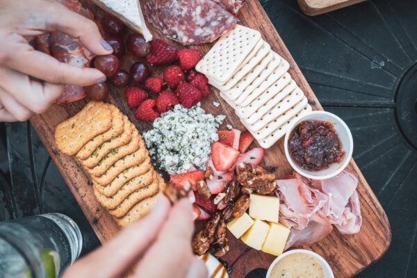 A close-up of a wooden charcuterie board with crackers, salami, prosciutto, cheese, grapes, raspberries, strawberries, herbed cheese, candied nuts, and small cups of jams or dips. Hands are reaching for food.