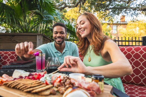 A smiling man and woman sit at an outdoor table, reaching for food on a charcuterie board with meats, cheeses, and crackers. Lush greenery surrounds them, and colorful drinks are on the table.