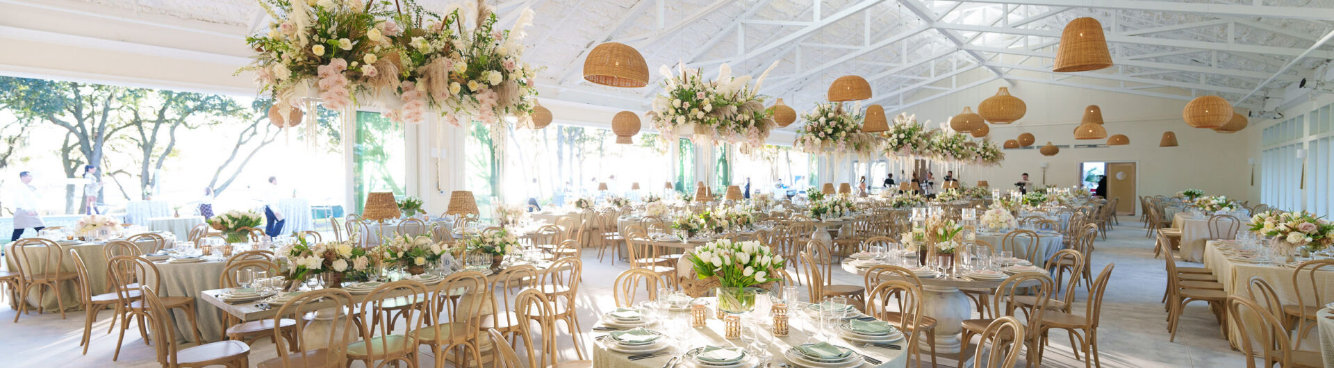 A bright, elegant banquet hall with round tables set for a formal event, decorated with white flowers, greenery, and hanging wicker lamps. Natural light streams through large windows.