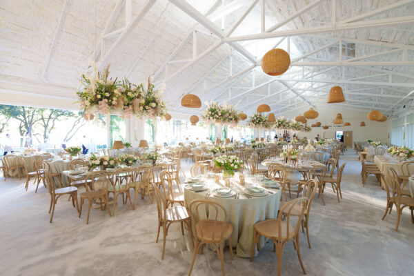 A bright, elegant banquet hall with round tables set for a formal event, decorated with white flowers, greenery, and hanging wicker lamps. Natural light streams through large windows.