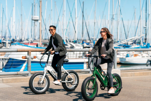 Two people ride electric bikes along a waterfront path on a sunny day, with numerous sailboats and masts visible in the marina behind them. Both are casually dressed and appear to be enjoying their ride.