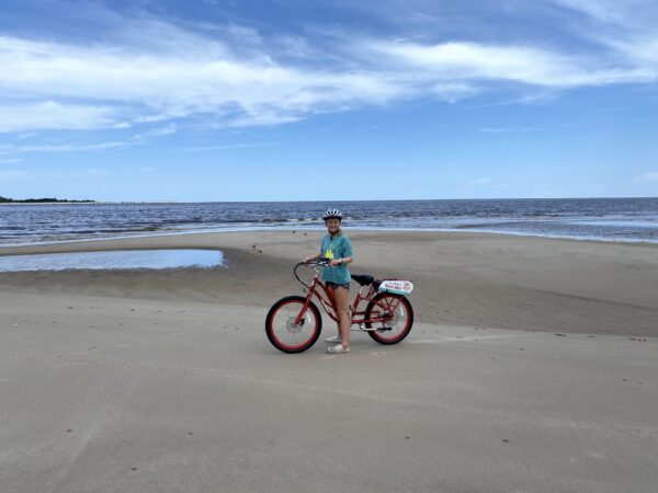 A person wearing a helmet and blue shirt stands next to a red bicycle on a sandy beach with water and blue sky in the background.