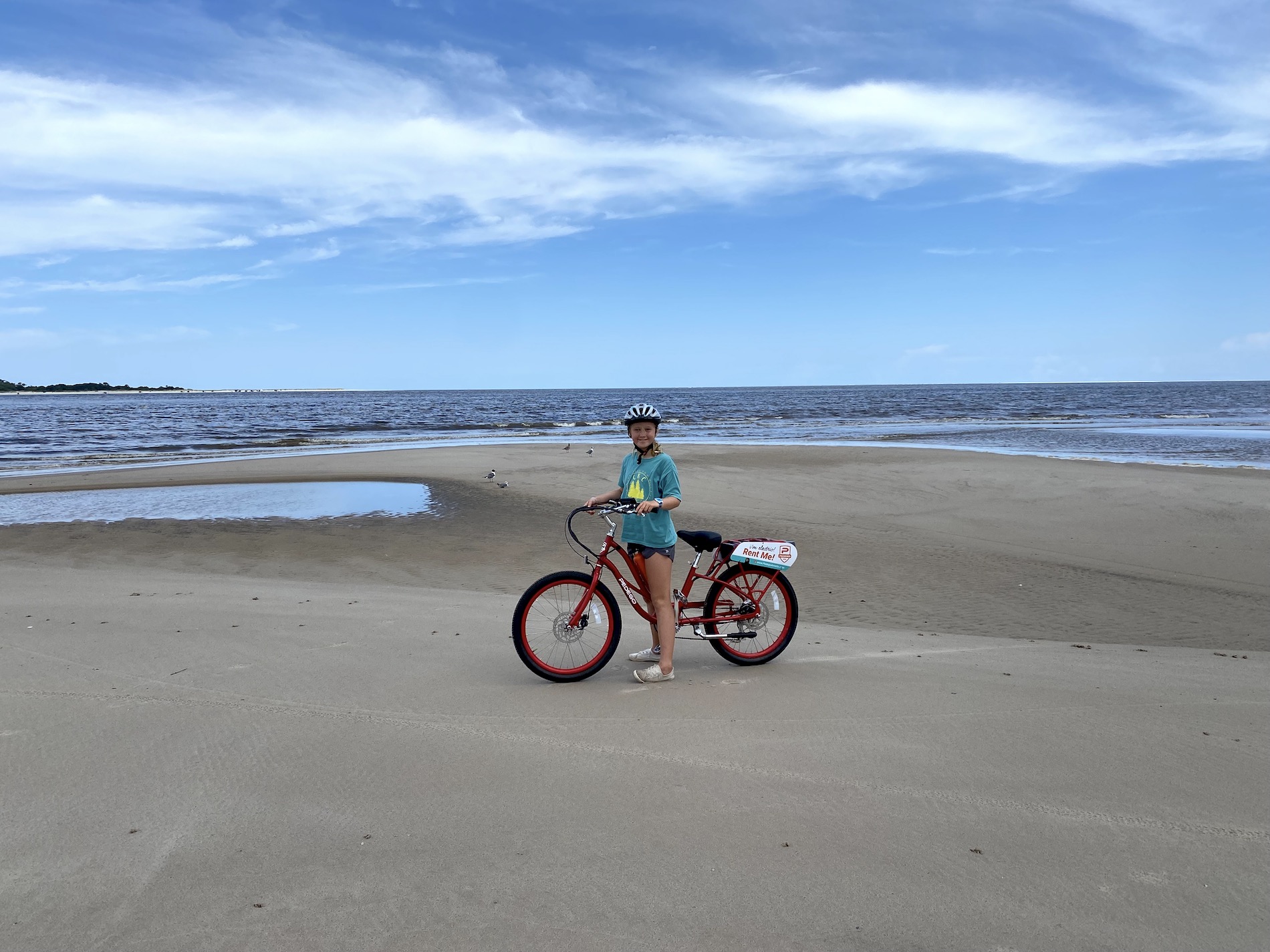 A person wearing a helmet and blue shirt stands next to a red bicycle on a sandy beach with water and blue sky in the background.