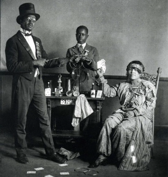 A vintage black-and-white photo shows three people performing a magic act: a man in a suit and top hat, a boy holding props, and a seated woman in a dress, holding playing cards, with props on a table behind them.