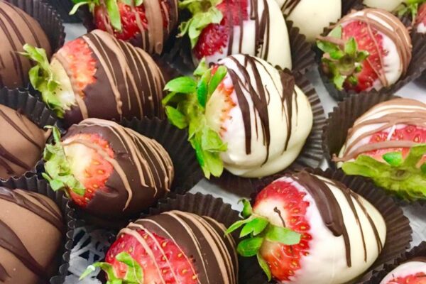 Close-up of strawberries dipped in milk, dark, and white chocolate, each drizzled with more chocolate, and arranged in black paper cups. The strawberries have green stems and appear fresh and glossy.