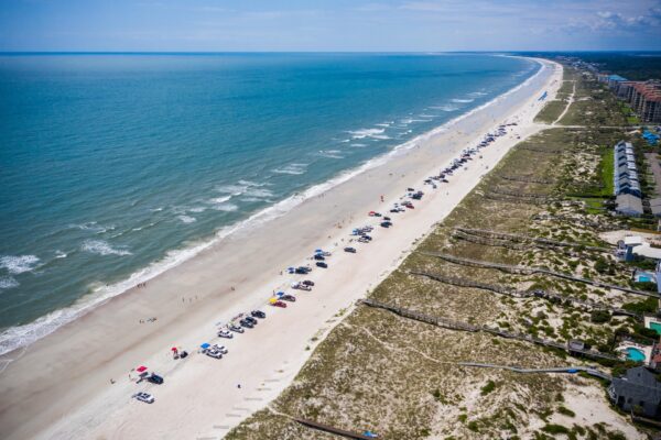 Aerial view of a long, sandy beach with cars and umbrellas lined up near the shore, waves gently rolling in, and rows of houses and greenery along the right side.