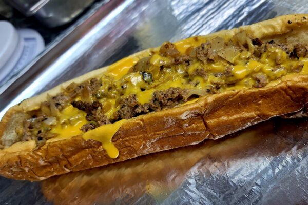 A close-up of a cheesesteak sandwich with melted cheddar cheese, ground beef, and grilled onions on a toasted hoagie roll, served on a sheet of aluminum foil.