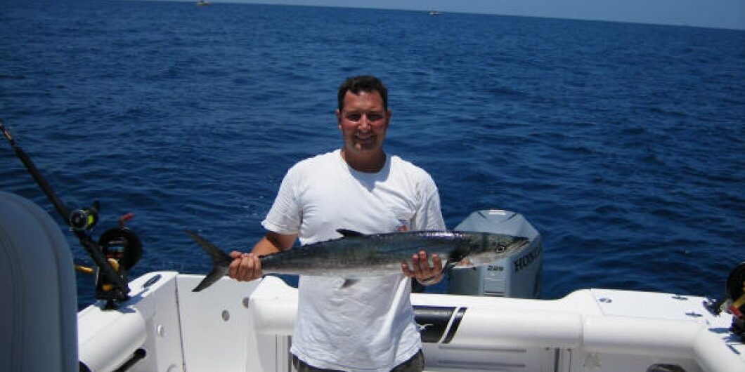A man standing on a boat holds a large fish, smiling at the camera. The boat is on open water under a clear blue sky, with another person seated nearby and fishing equipment visible.
