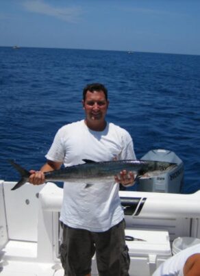 A man standing on a boat holds a large fish, smiling at the camera. The boat is on open water under a clear blue sky, with another person seated nearby and fishing equipment visible.