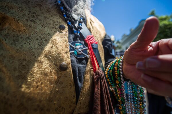 A person wearing a gold-patterned vest and multiple colorful beaded necklaces, including a pirate skull pendant, gives a thumbs up outdoors on a sunny day.