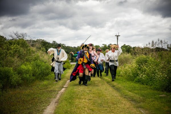 A group of people dressed as pirates, led by a person in a colorful pirate costume with a hat and sword, walk energetically down a grassy path surrounded by greenery under a cloudy sky.