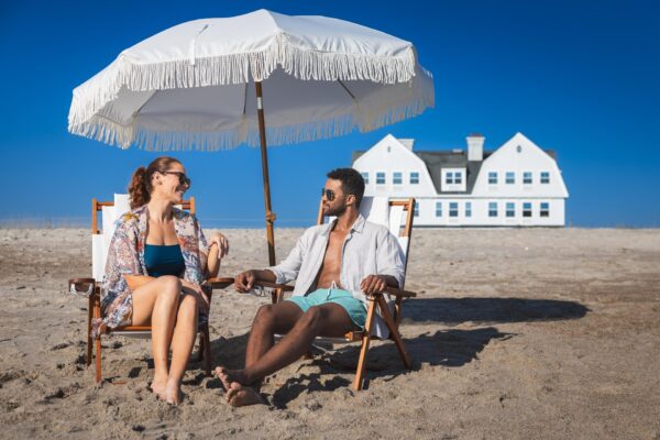 A smiling man and woman sit in beach chairs under a white umbrella on a sandy beach, wearing summer clothes and sunglasses, with a large white house in the background against a clear blue sky.