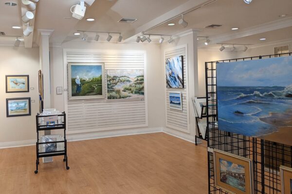 Art gallery interior with landscape and seascape paintings on white walls and black grid panels, spotlights overhead, wood flooring, and a small display cart with art prints on the left side.