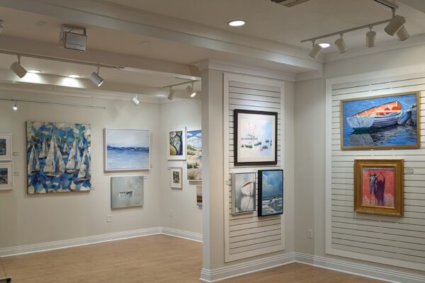 Art gallery with wooden floors, white walls, and track lighting displays paintings of boats, seascapes, and landscapes in various frames arranged neatly on the walls.