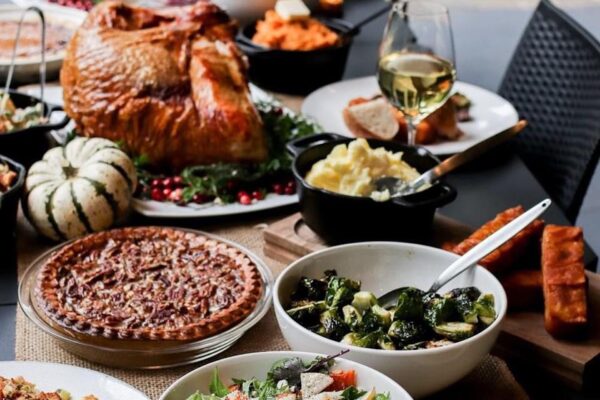 A festive table is set with a roast turkey, pecan pie, mashed potatoes, Brussels sprouts, salad, bread, sweet potatoes, a small decorative white pumpkin, and a glass of white wine.