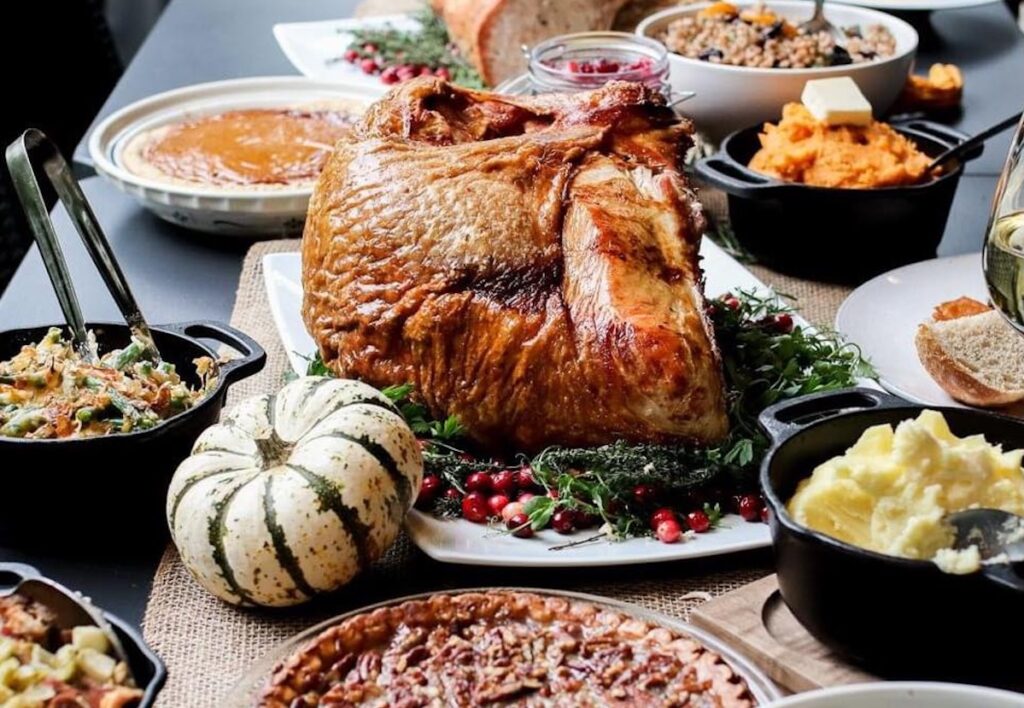 A Thanksgiving feast featuring a roasted turkey, mashed potatoes, gravy, pumpkin pie, pecan pie, sweet potatoes, cranberry sauce, stuffing, bread, coleslaw, and a small decorative pumpkin.
