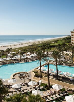 A beachfront resort with a large pool, lounge chairs, palm trees, and a multi-story hotel overlooks a sandy beach and the ocean under a clear blue sky.