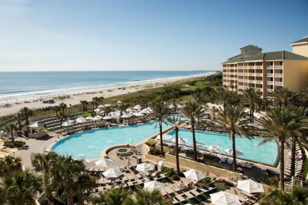 A beachfront resort with a large pool, lounge chairs, palm trees, and a multi-story hotel overlooks a sandy beach and the ocean under a clear blue sky.