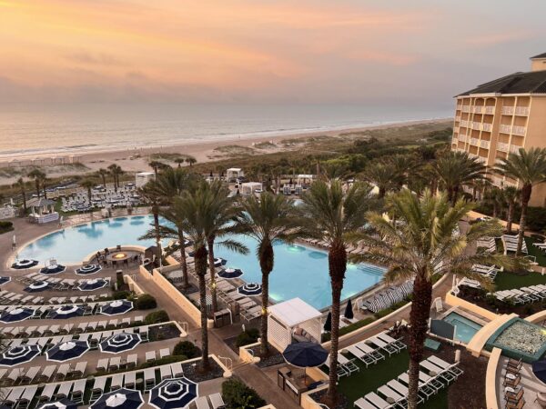 A beach resort at sunset with two pools, lounge chairs, palm trees, and a sandy beach in the background. The sky is orange and pink, creating a peaceful atmosphere.