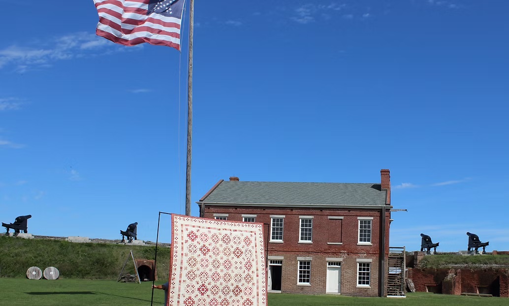 A large American flag waves in front of a historic red brick building, with cannons lined up on a grassy fort wall in the background and a patterned quilt hanging on display in the foreground under a clear blue sky.