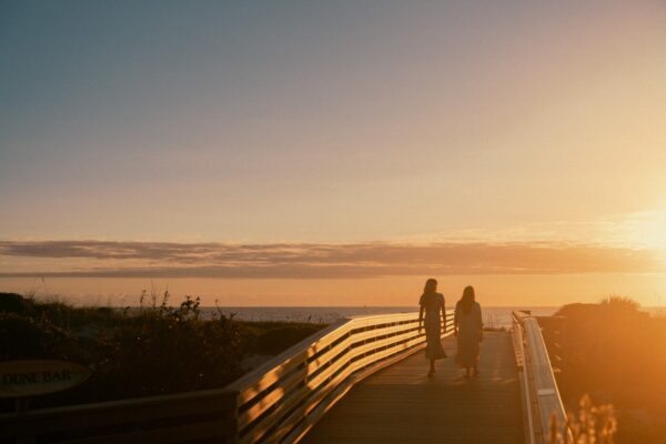 Two people walk along a wooden boardwalk toward the beach at sunset, with a golden sky and the sun low on the horizon. Vegetation lines both sides of the boardwalk.