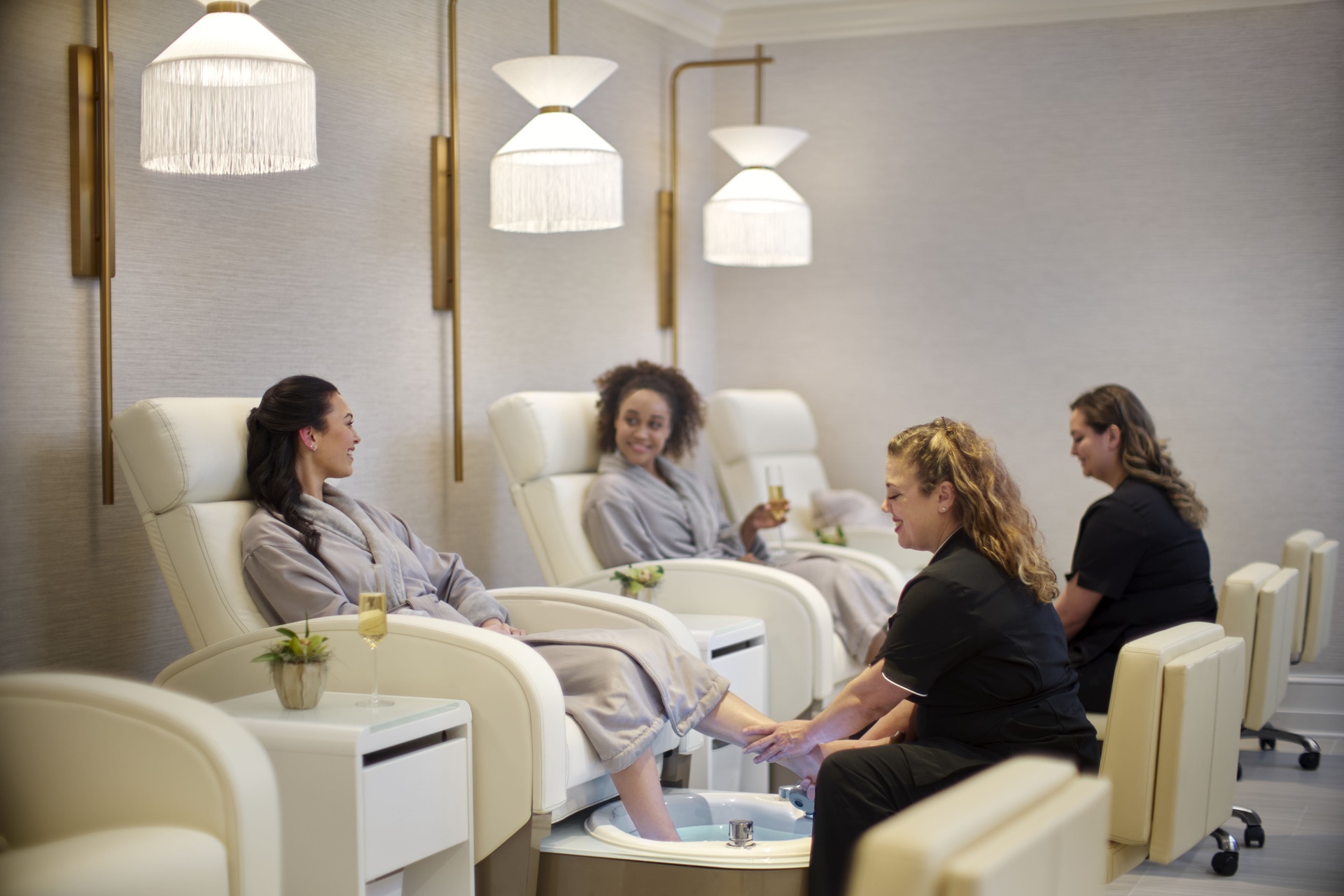Two women in robes relax in spa chairs, smiling and chatting, while spa staff provide pedicure treatments. Soft lighting and elegant decor create a calm, luxurious atmosphere.