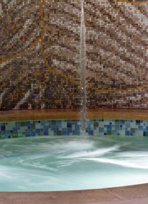 A small, round indoor spa pool with bubbling water, tiled edge, and mosaic tile wall. A cushioned chair and side table with a plant sit nearby on the light-colored tile floor.