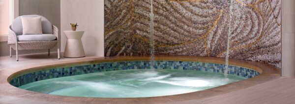 A small, round indoor spa pool with bubbling water, tiled edge, and mosaic tile wall. A cushioned chair and side table with a plant sit nearby on the light-colored tile floor.