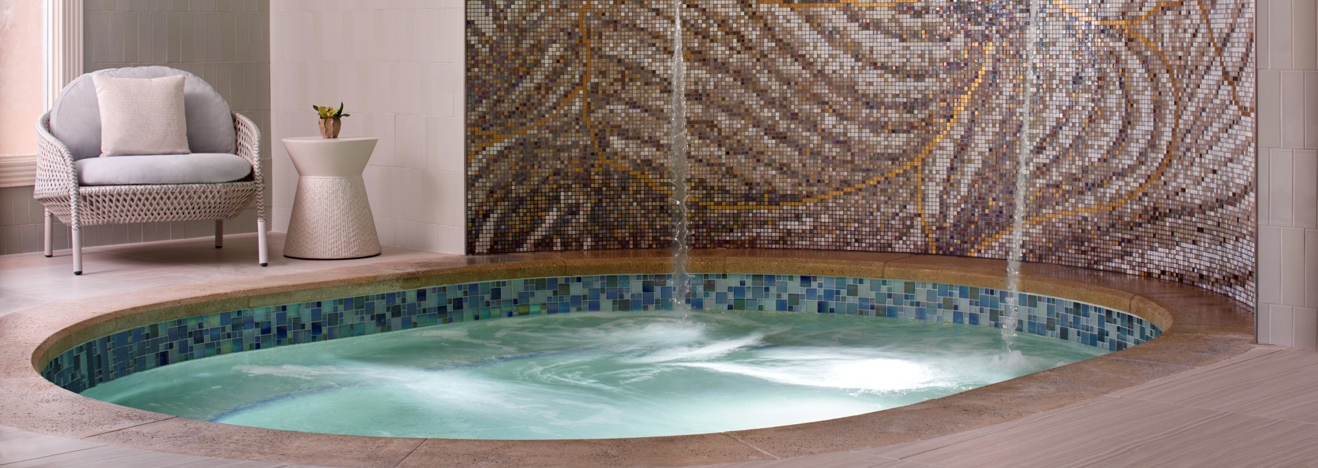 A small, round indoor spa pool with bubbling water, tiled edge, and mosaic tile wall. A cushioned chair and side table with a plant sit nearby on the light-colored tile floor.