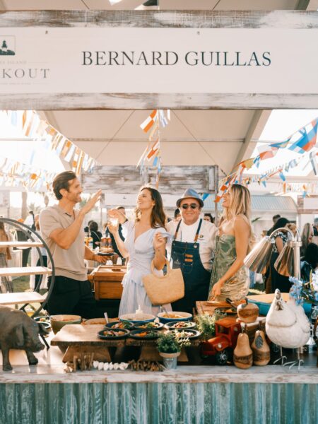 Four people smile and enjoy drinks at an outdoor food booth labeled Bernard Guillas, decorated with colorful streamers and food. The booth features various dishes, wooden decor, and a small stuffed animal.