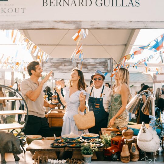 Four people smile and enjoy drinks at an outdoor food booth labeled Bernard Guillas, decorated with colorful streamers and food. The booth features various dishes, wooden decor, and a small stuffed animal.