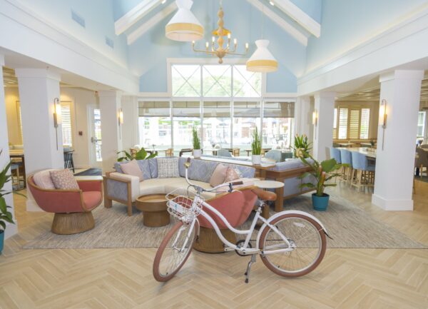A bright, spacious lounge with pastel furniture, indoor plants, wooden tables, and a white bicycle with a basket in the center. Large windows let in natural light and a chandelier hangs from the vaulted ceiling.