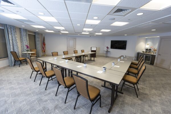 Conference room with U-shaped table arrangement, brown chairs, notepads, cups, and pens on tables, an American flag in the corner, a large flat screen TV, and a refreshment station on the right.