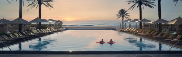 Two people relax in a large, tranquil swimming pool at sunset, surrounded by lounge chairs, umbrellas, and palm trees, with the ocean visible in the background.