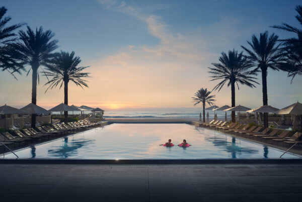 Two people swim in a large, rectangular pool surrounded by lounge chairs and palm trees at sunset, with the ocean visible in the background and a clear sky overhead.