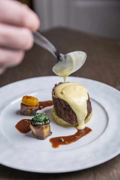 A hand holds a spoon, pouring creamy yellow sauce over a piece of beef atop mashed potatoes, with two small garnished vegetables and a drizzle of brown sauce on a white plate.