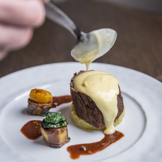 A hand holds a spoon, pouring creamy yellow sauce over a piece of beef atop mashed potatoes, with two small garnished vegetables and a drizzle of brown sauce on a white plate.