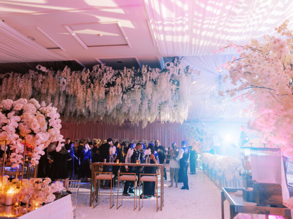 A lavish indoor wedding reception with guests mingling under hanging white floral arrangements. Elegant tables are decorated with white flowers, and soft pink and purple lighting fills the room.