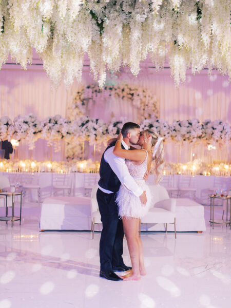 A newlywed couple shares a romantic dance under a ceiling of hanging white flowers in an elegant, flower-filled wedding venue glowing with soft, warm lights.
