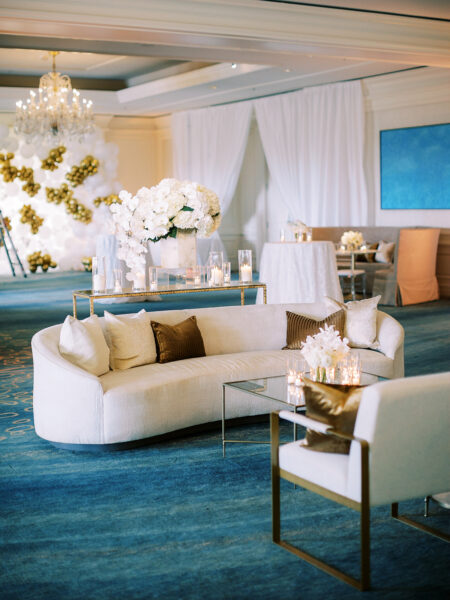 Elegant event space with white curved sofas, glass tables, gold and white decorations, and a large floral centerpiece. A balloon arch and chandelier add a luxurious touch. The floor is covered with a rich blue carpet.